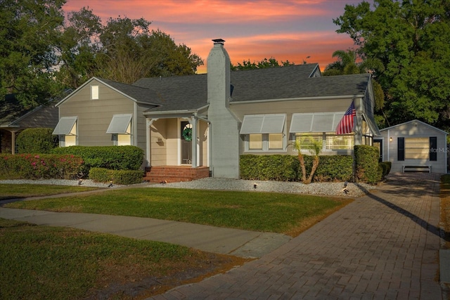 view of front facade featuring decorative driveway, a chimney, a front yard, an outbuilding, and a shingled roof