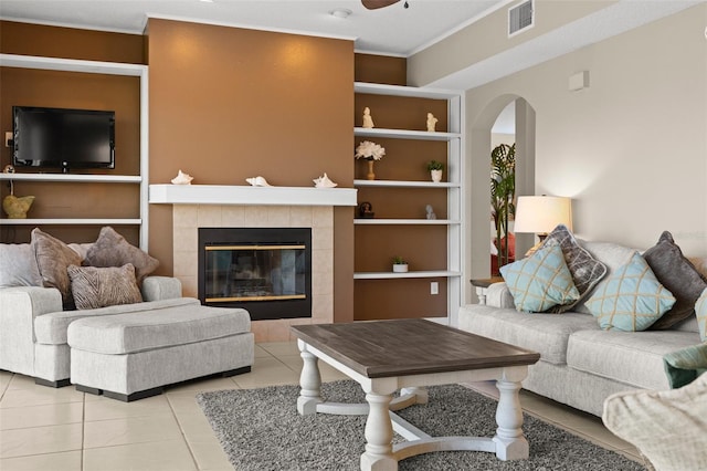 living area featuring built in shelves, light tile patterned flooring, arched walkways, a tiled fireplace, and crown molding