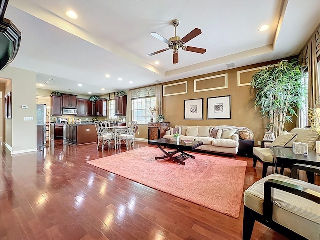living room with a raised ceiling, dark wood-type flooring, and ceiling fan