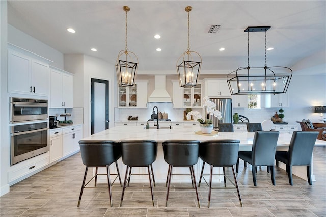 kitchen with glass insert cabinets, white cabinetry, custom exhaust hood, appliances with stainless steel finishes, and a spacious island