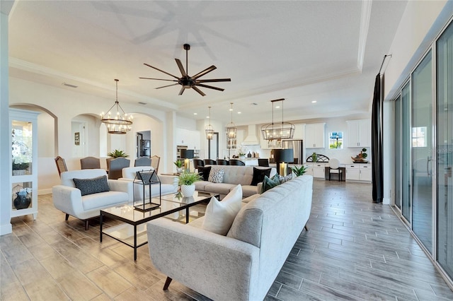 living area featuring crown molding, a tray ceiling, wood tiled floor, arched walkways, and ceiling fan with notable chandelier