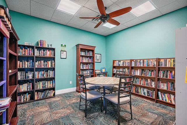 carpeted office with a ceiling fan, wall of books, baseboards, and a drop ceiling