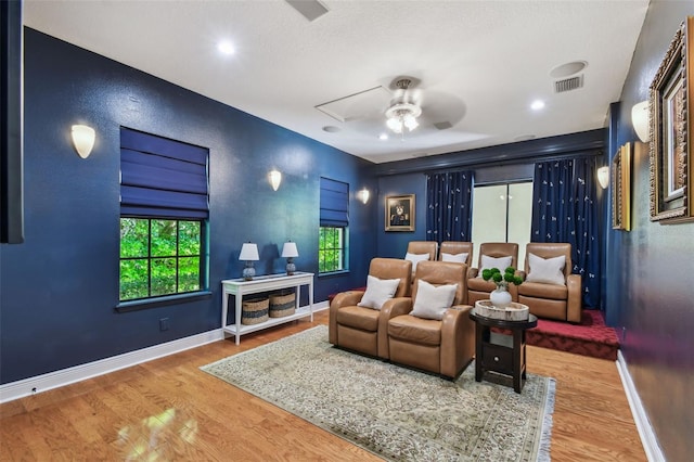 home theater featuring wood finished floors, a ceiling fan, visible vents, baseboards, and a textured wall