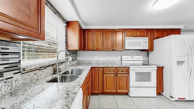 kitchen with brown cabinets, a sink, white appliances, light tile patterned floors, and light stone countertops