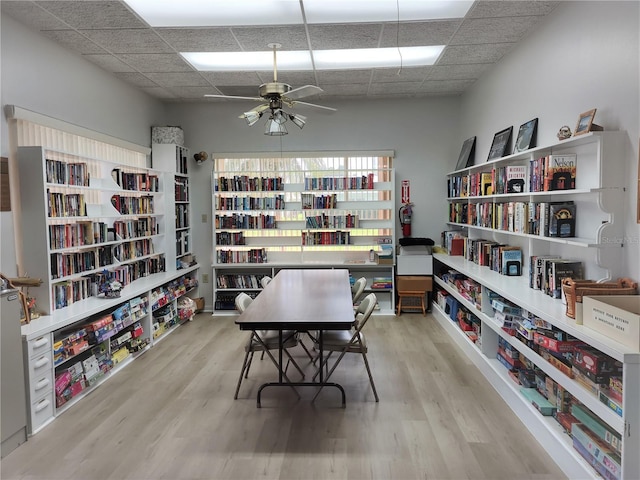 home office with a drop ceiling, wall of books, ceiling fan, and wood finished floors