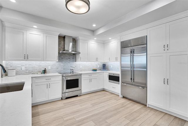 kitchen featuring wall chimney range hood, a sink, light wood-type flooring, white cabinets, and appliances with stainless steel finishes