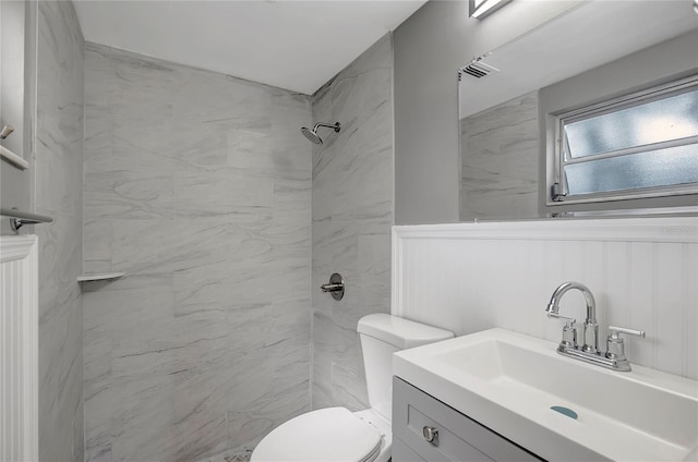 full bathroom featuring toilet, a wainscoted wall, tiled shower, and vanity