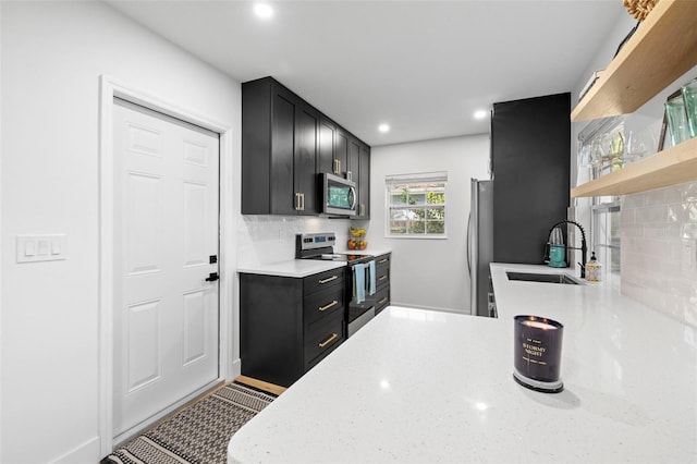 kitchen featuring stainless steel appliances, a sink, open shelves, dark cabinetry, and light stone counters