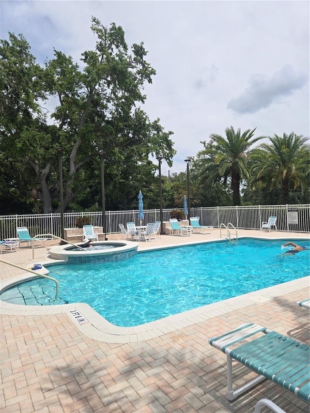 community pool with a patio, fence, a hot tub, and view of scattered trees