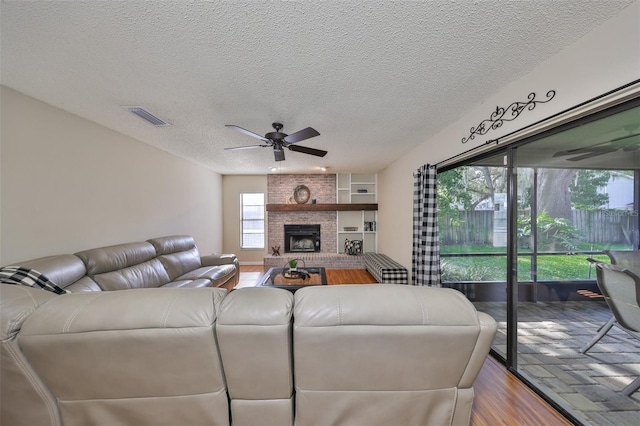 living room with wood finished floors, a textured ceiling, a brick fireplace, visible vents, and ceiling fan