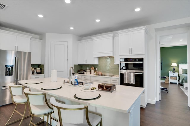 kitchen featuring dark wood finished floors, an island with sink, a sink, high end refrigerator, and multiple ovens