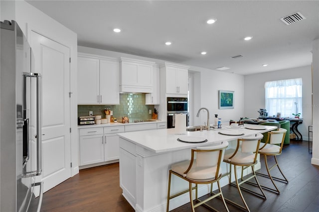 kitchen featuring an island with sink, stainless steel appliances, visible vents, decorative backsplash, and dark wood-style flooring