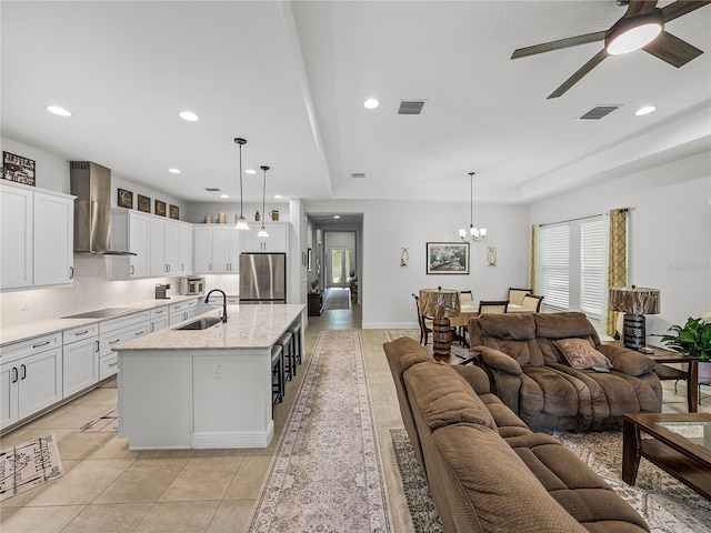 kitchen with stainless steel refrigerator, black electric stovetop, ceiling fan with notable chandelier, open floor plan, and wall chimney range hood