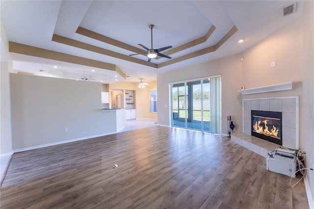 unfurnished living room with a tile fireplace, a raised ceiling, wood finished floors, baseboards, and ceiling fan