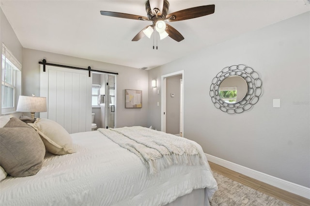 bedroom with baseboards, a barn door, light wood finished floors, ceiling fan, and ensuite bathroom