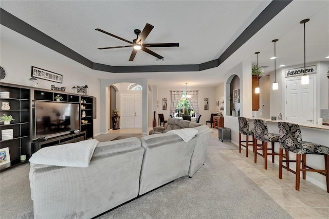 living area with baseboards, a raised ceiling, arched walkways, light colored carpet, and ceiling fan with notable chandelier