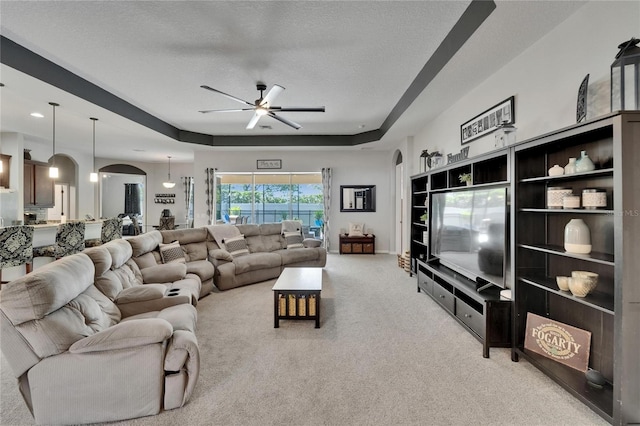 living area featuring a tray ceiling, arched walkways, a textured ceiling, light colored carpet, and a ceiling fan