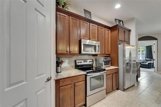 kitchen featuring light stone counters, light tile patterned floors, brown cabinetry, decorative backsplash, and stainless steel appliances