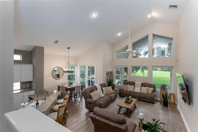 living area featuring ceiling fan with notable chandelier, high vaulted ceiling, baseboards, light wood finished floors, and visible vents