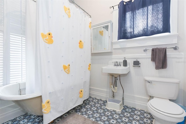 full bath with a wainscoted wall, tile patterned floors, a shower with shower curtain, toilet, and a soaking tub