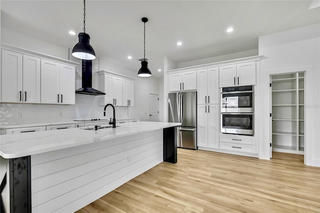 kitchen with a sink, wall chimney exhaust hood, appliances with stainless steel finishes, white cabinets, and light wood-style floors