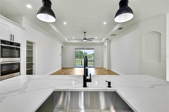 kitchen featuring a sink, a raised ceiling, light wood finished floors, stainless steel double oven, and hanging light fixtures