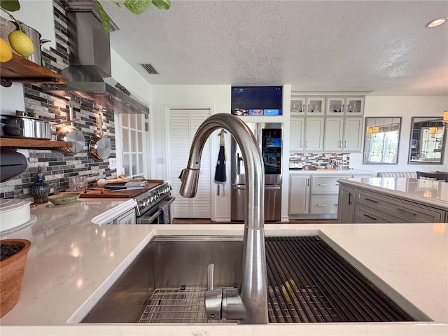 kitchen with decorative backsplash, a textured ceiling, wall chimney exhaust hood, white cabinetry, and glass insert cabinets