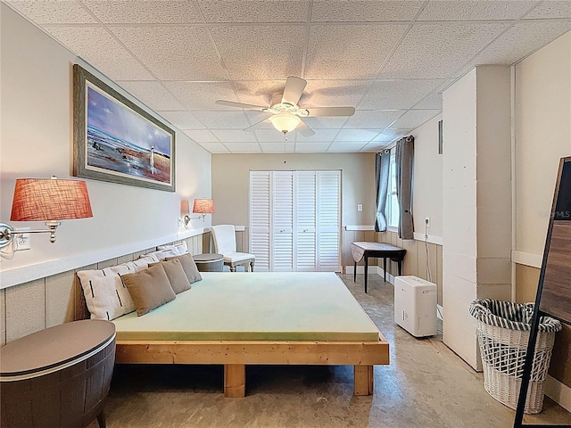 bedroom featuring a drop ceiling, concrete floors, wainscoting, and a ceiling fan