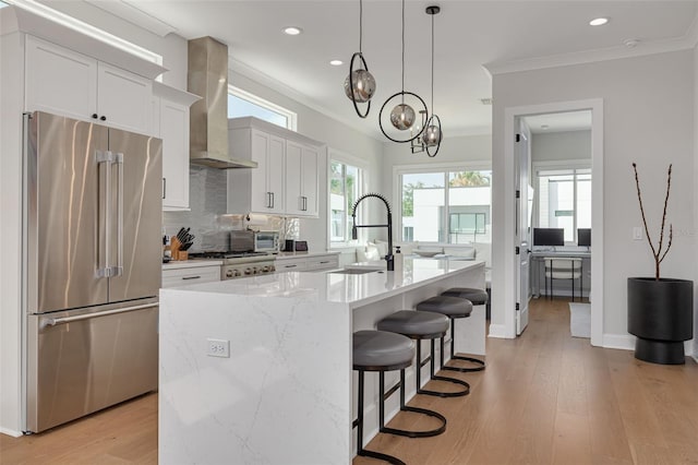 kitchen featuring ornamental molding, a sink, wall chimney exhaust hood, a center island with sink, and high end refrigerator