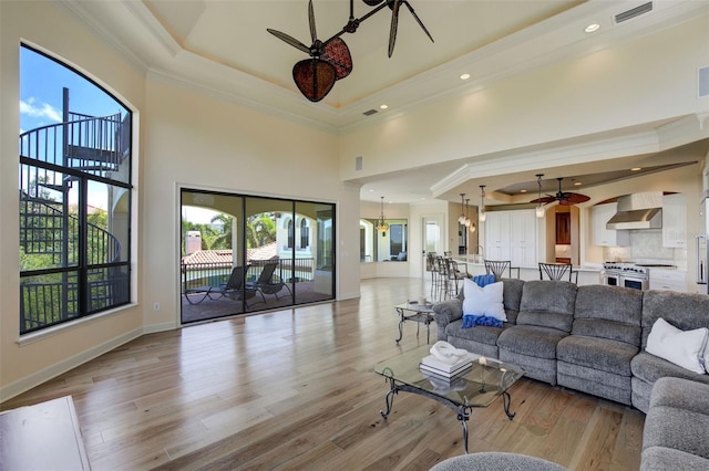 living room with crown molding, ceiling fan with notable chandelier, baseboards, a raised ceiling, and light wood finished floors