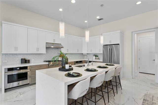 kitchen featuring decorative backsplash, marble finish floor, stainless steel appliances, under cabinet range hood, and a kitchen breakfast bar
