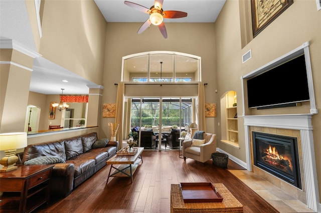 living room featuring visible vents, a tiled fireplace, ceiling fan with notable chandelier, hardwood / wood-style floors, and a high ceiling