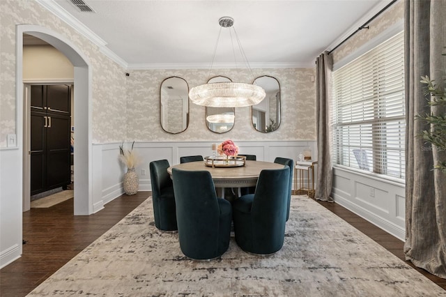 dining space with wallpapered walls, crown molding, a wainscoted wall, dark wood-type flooring, and arched walkways