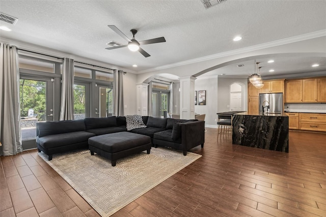 living area featuring arched walkways, ornate columns, a ceiling fan, visible vents, and dark wood-style floors