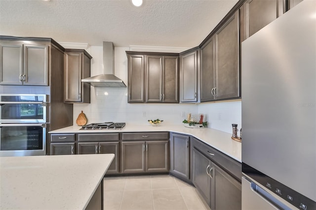 kitchen with appliances with stainless steel finishes, dark brown cabinetry, wall chimney range hood, backsplash, and light tile patterned floors