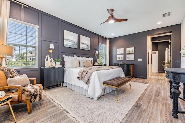 bedroom with a decorative wall, visible vents, light wood-style floors, and multiple windows