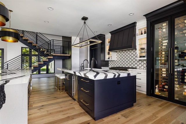 kitchen with glass insert cabinets, custom range hood, tasteful backsplash, pendant lighting, and an island with sink