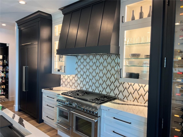 kitchen with double oven range, decorative backsplash, light wood-style flooring, custom exhaust hood, and dark cabinetry