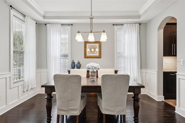 dining area featuring a decorative wall, a raised ceiling, arched walkways, and dark wood finished floors