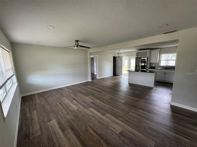 unfurnished living room with ceiling fan with notable chandelier, dark wood-type flooring, a textured ceiling, visible vents, and baseboards