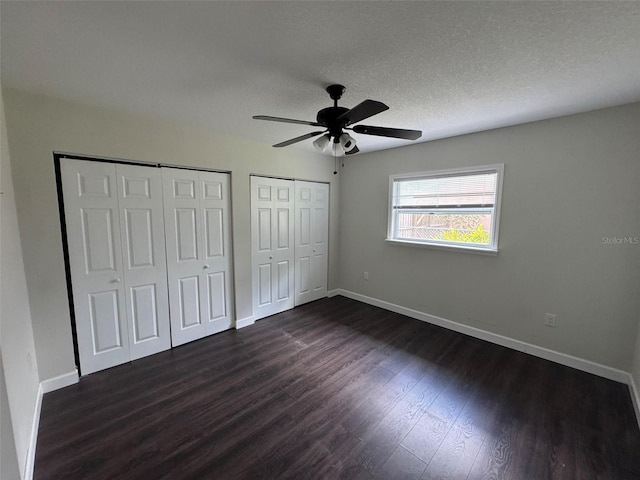 unfurnished bedroom featuring ceiling fan, a textured ceiling, multiple closets, baseboards, and dark wood-style floors