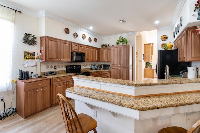 kitchen featuring black appliances, light wood finished floors, a kitchen bar, light stone counters, and brown cabinets