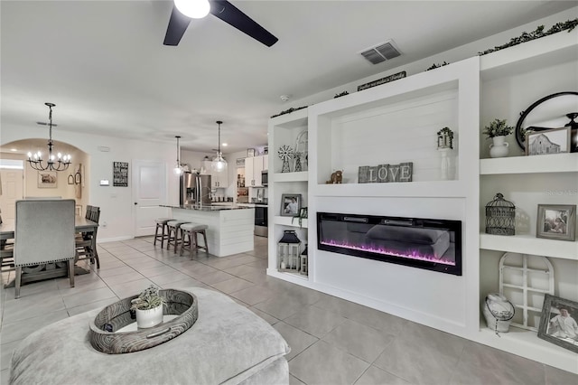 living room featuring light tile patterned floors, a glass covered fireplace, visible vents, and ceiling fan with notable chandelier
