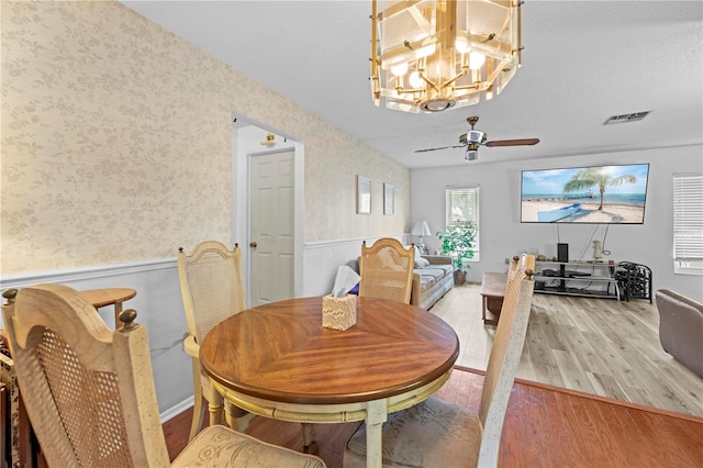 dining space with visible vents, wood finished floors, a wainscoted wall, ceiling fan with notable chandelier, and wallpapered walls