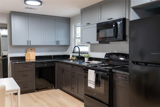 kitchen with a sink, light wood-type flooring, black appliances, dark stone counters, and gray cabinets