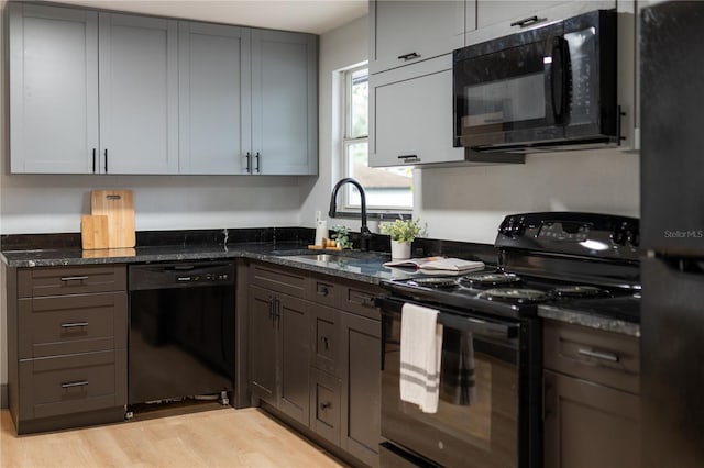 kitchen featuring a sink, light wood-type flooring, black appliances, gray cabinetry, and dark stone counters