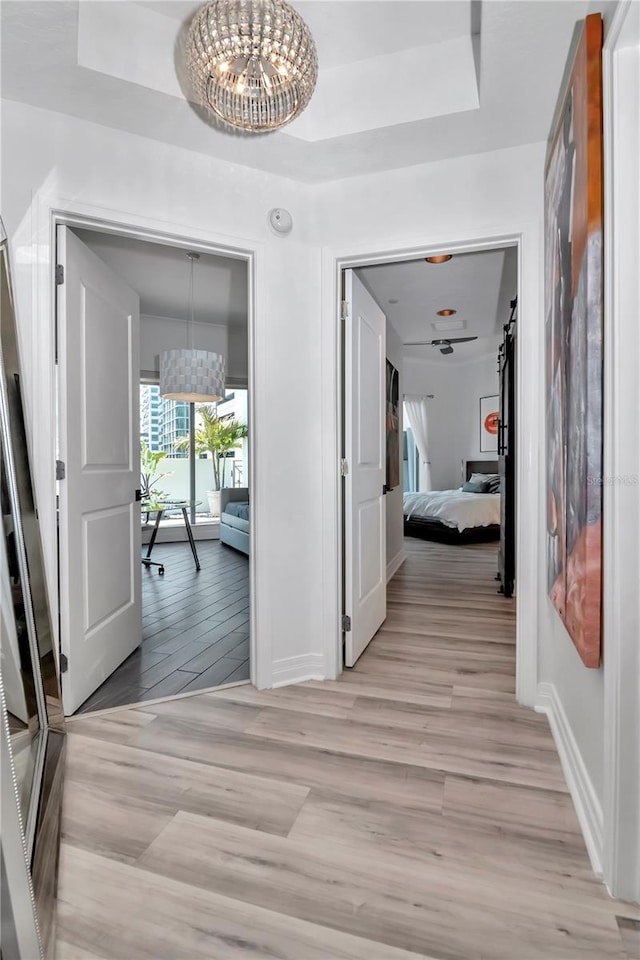 hall featuring light wood finished floors, baseboards, a chandelier, and a tray ceiling