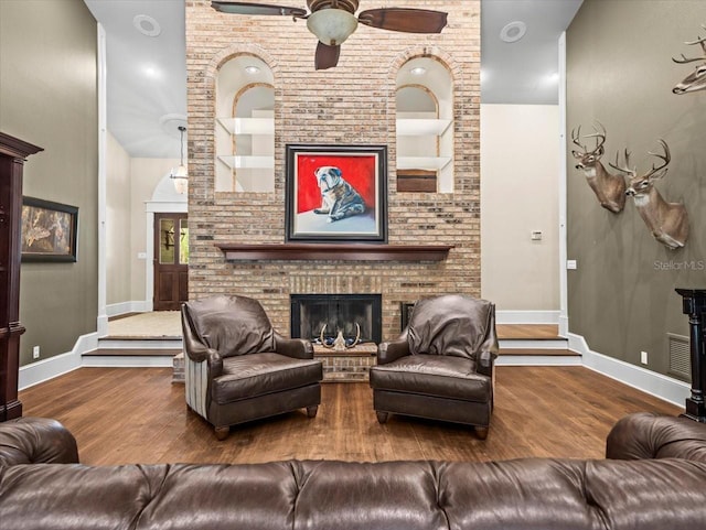 living area featuring wood finished floors, built in shelves, ceiling fan, a brick fireplace, and baseboards