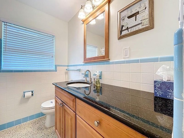 bathroom featuring speckled floor, vanity, toilet, wainscoting, and tile walls