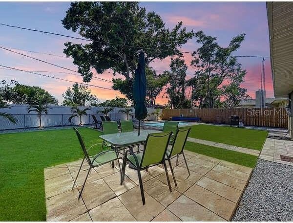 patio terrace at dusk with a fenced backyard, outdoor dining space, a lawn, and a pool